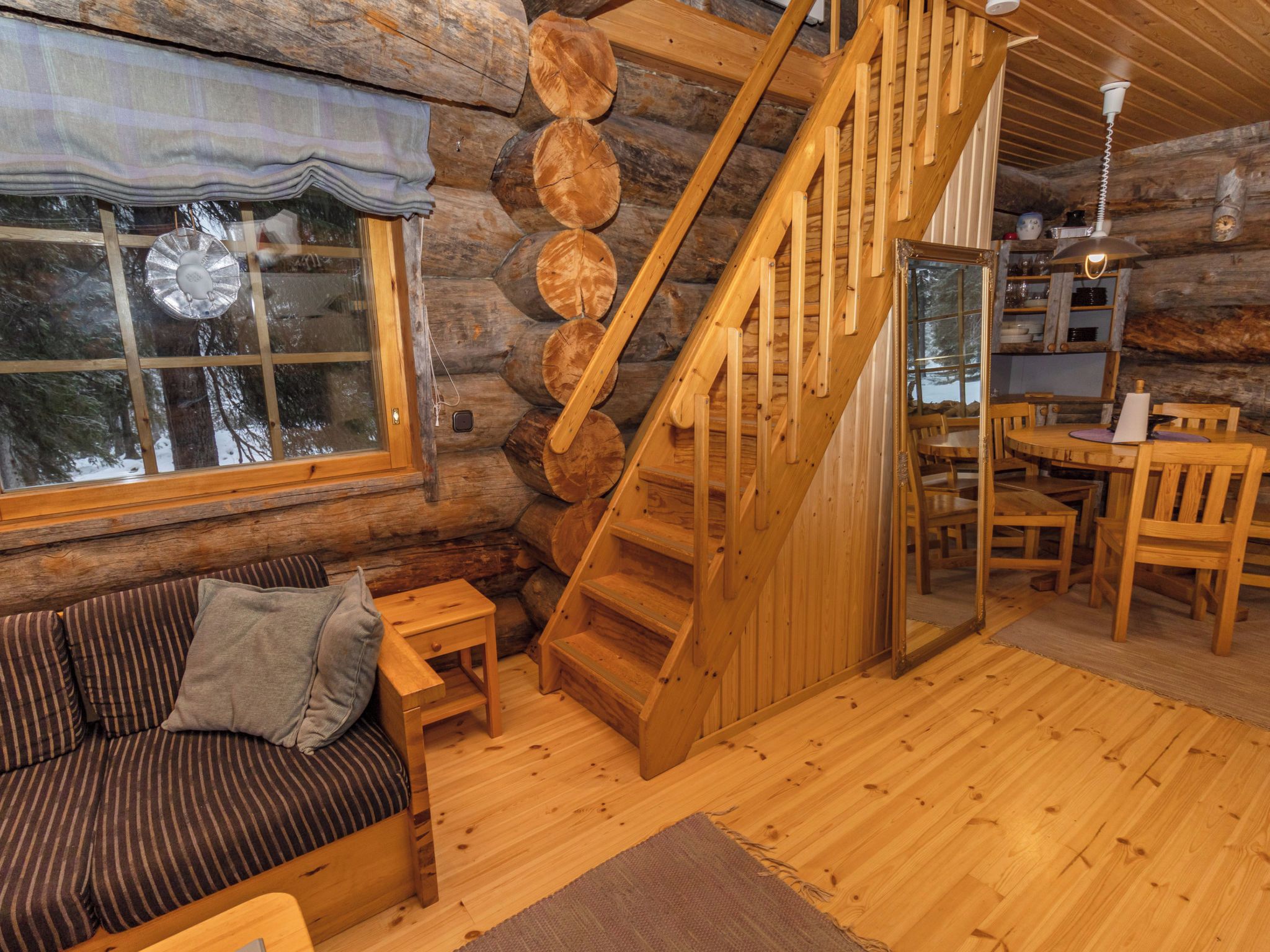 Interior de una cabaña de troncos con una escalera de madera, un sofá, una mesa de comedor y una ventana con vista a árboles.