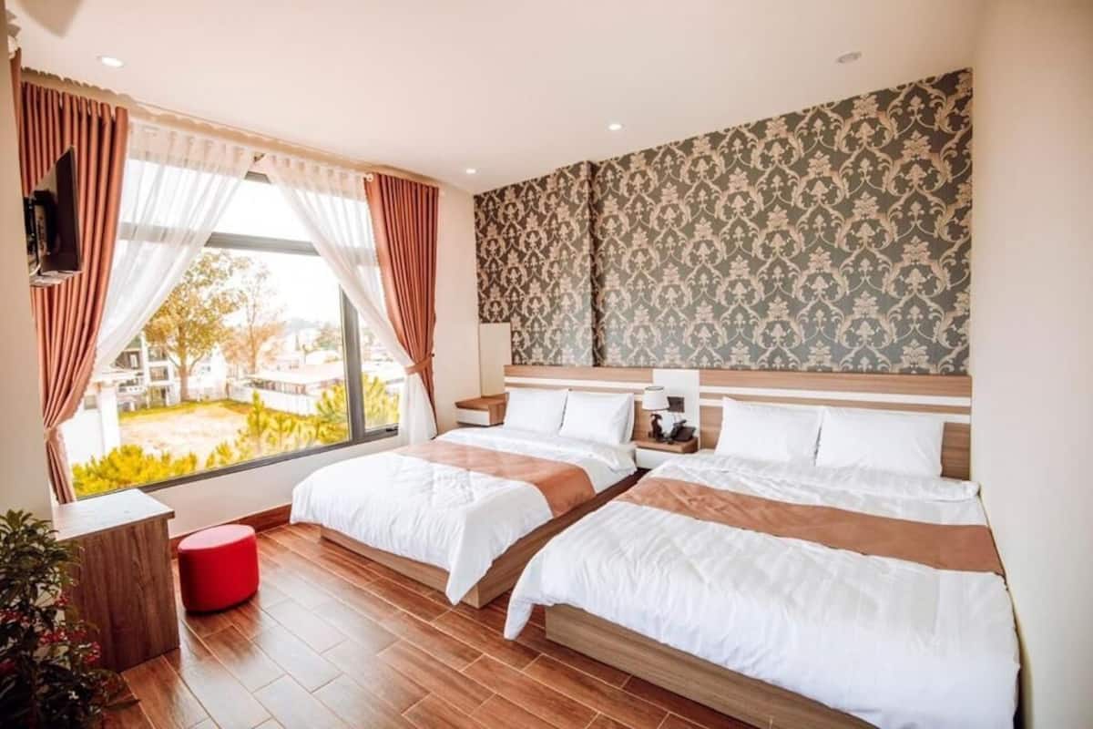 A hotel room with two beds, a large window with curtains, a wooden floor, and a patterned wallpaper.
