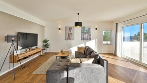 Un salon spacieux doté d’un grand téléviseur à écran plat, d’un canapé d’angle gris, d’une table basse en bois et d’un lampadaire. Deux œuvres d’art sont accrochées au mur, et une fenêtre offre une vue sur la verdure.