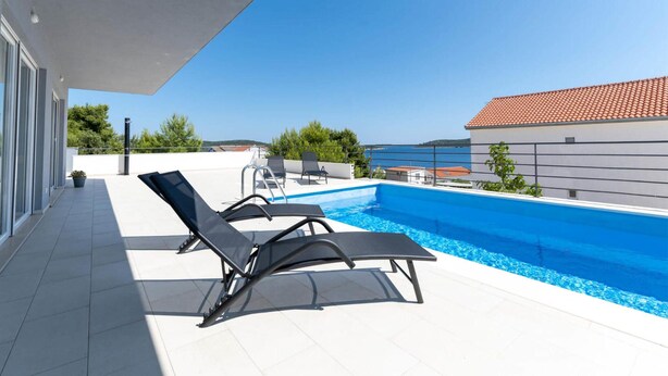 Une piscine sur le toit, avec deux chaises longues, une vue dégagée sur la mer et, au loin, un bâtiment dont le toit est recouvert de tuiles.