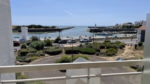 Vista costera desde un balcón con vista a un puerto deportivo con barcos, un faro y una carretera con coches.