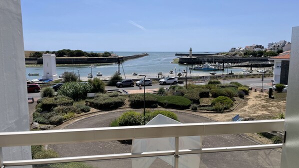 A coastal view from a balcony overlooking a marina with boats, a lighthouse, and a road with cars.
