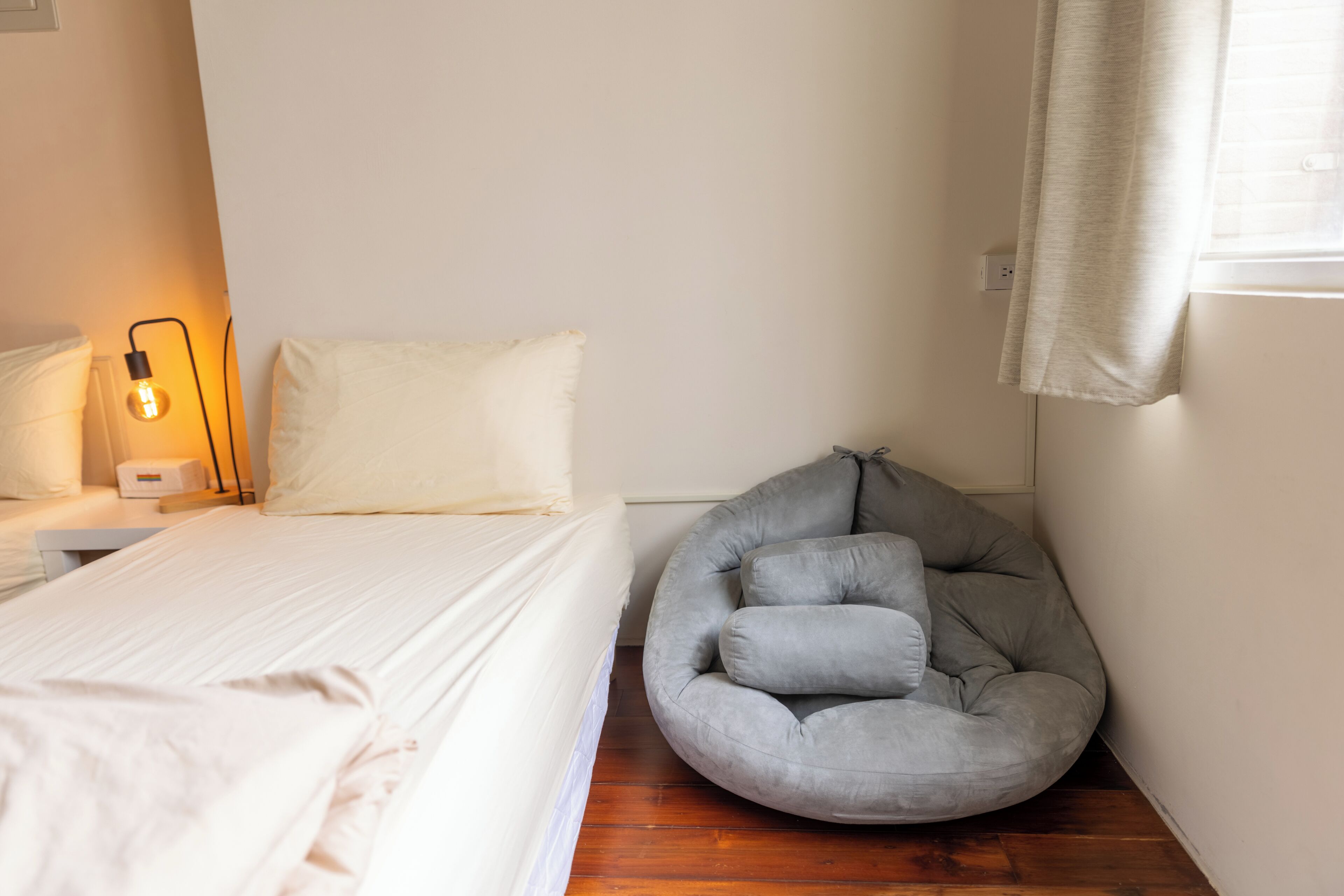 A bedroom with a bed, a bedside lamp, a grey bean bag chair, and a window with curtains.