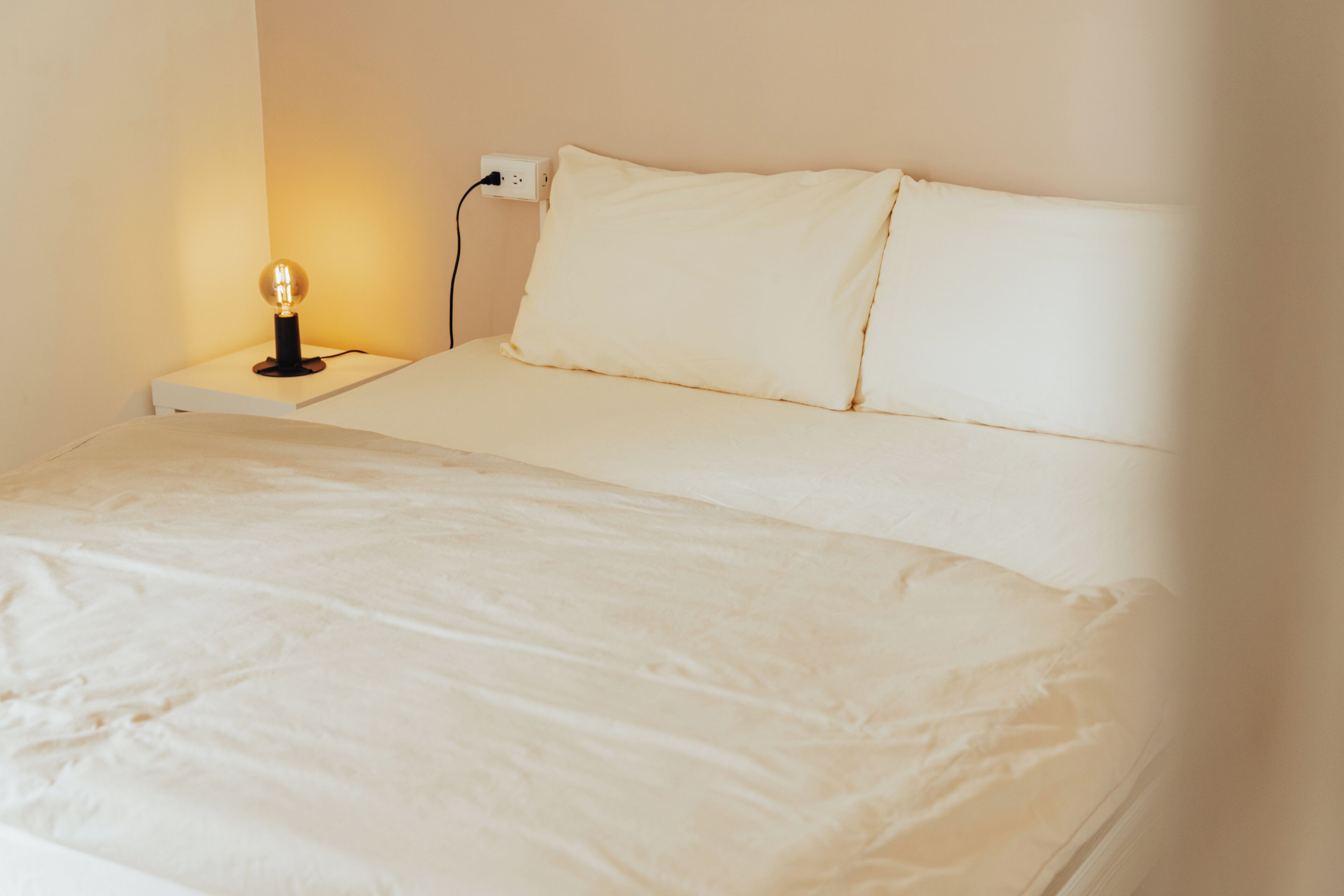 A neatly made bed with white linens, a bedside lamp, and a power outlet on the wall.