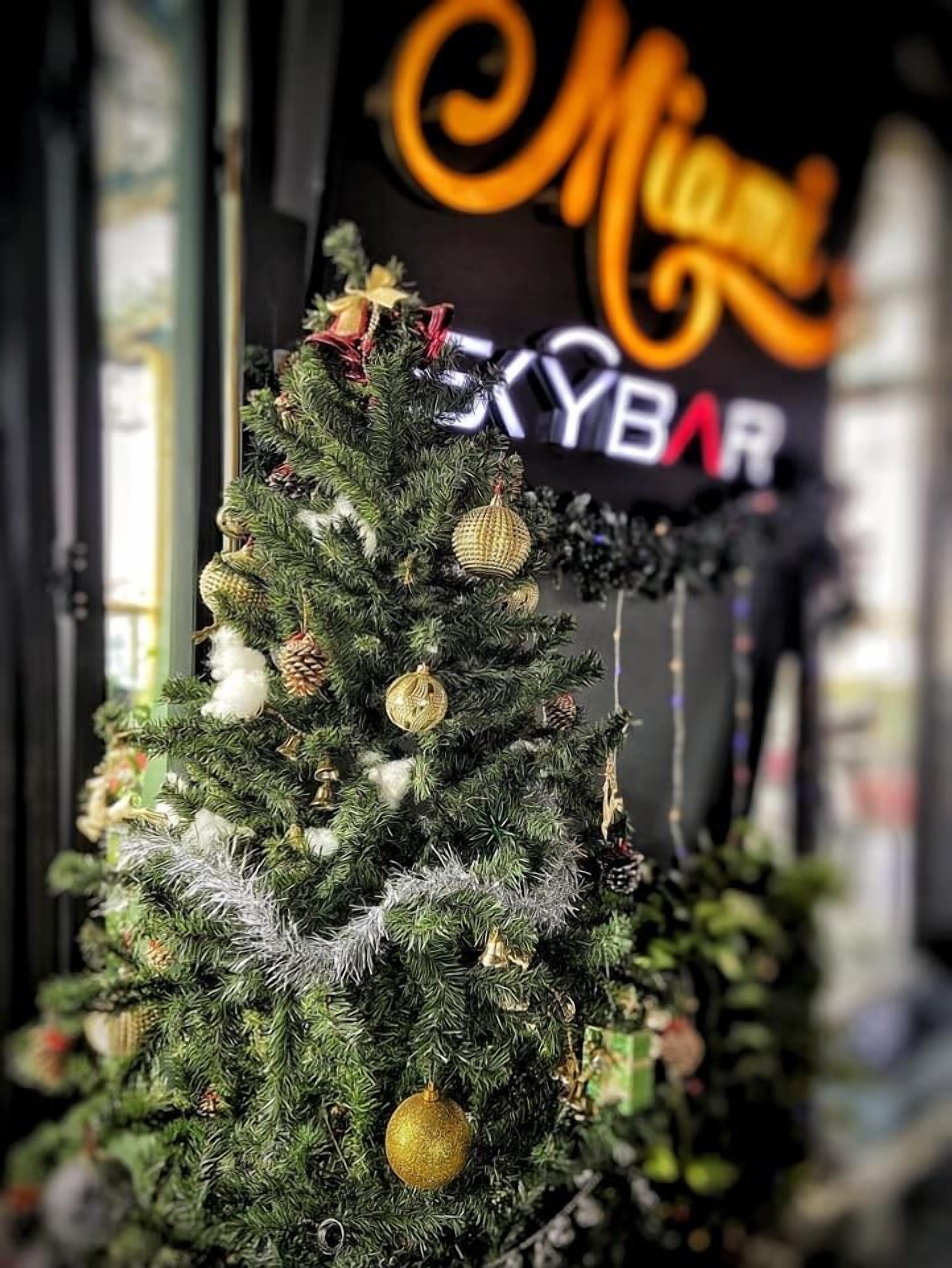 A Christmas tree decorated with ornaments and pine cones, with a sign reading "Mia's Skybar" in the background.