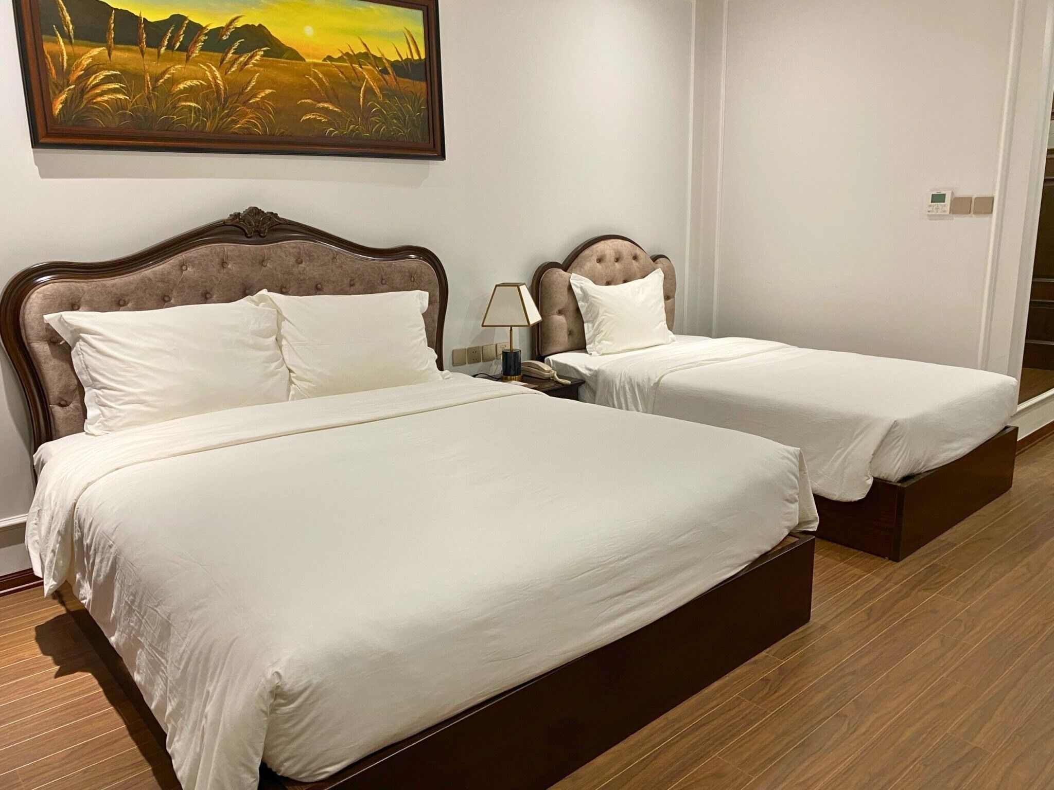A hotel room with two beds, a nightstand with a lamp, a painting on the wall, and a wooden floor.