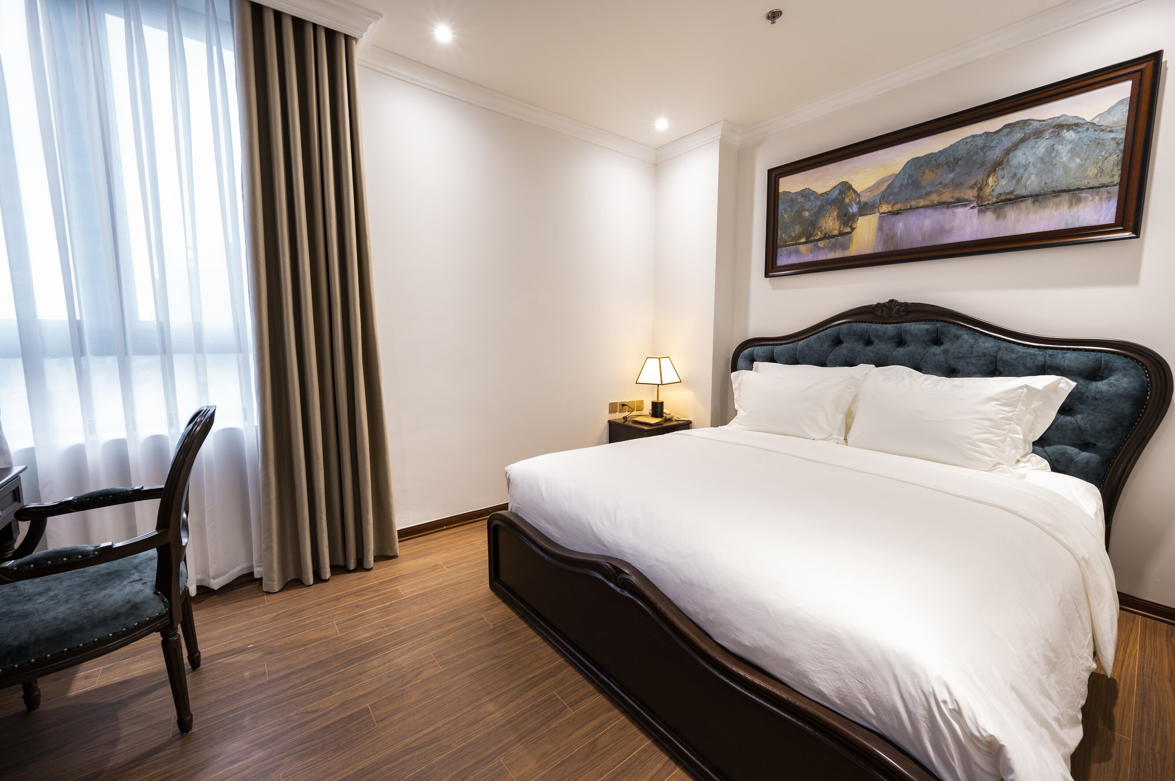 Habitación de hotel ordenada con una cama grande, una silla, una mesita de noche con lámpara y un cuadro en la pared.