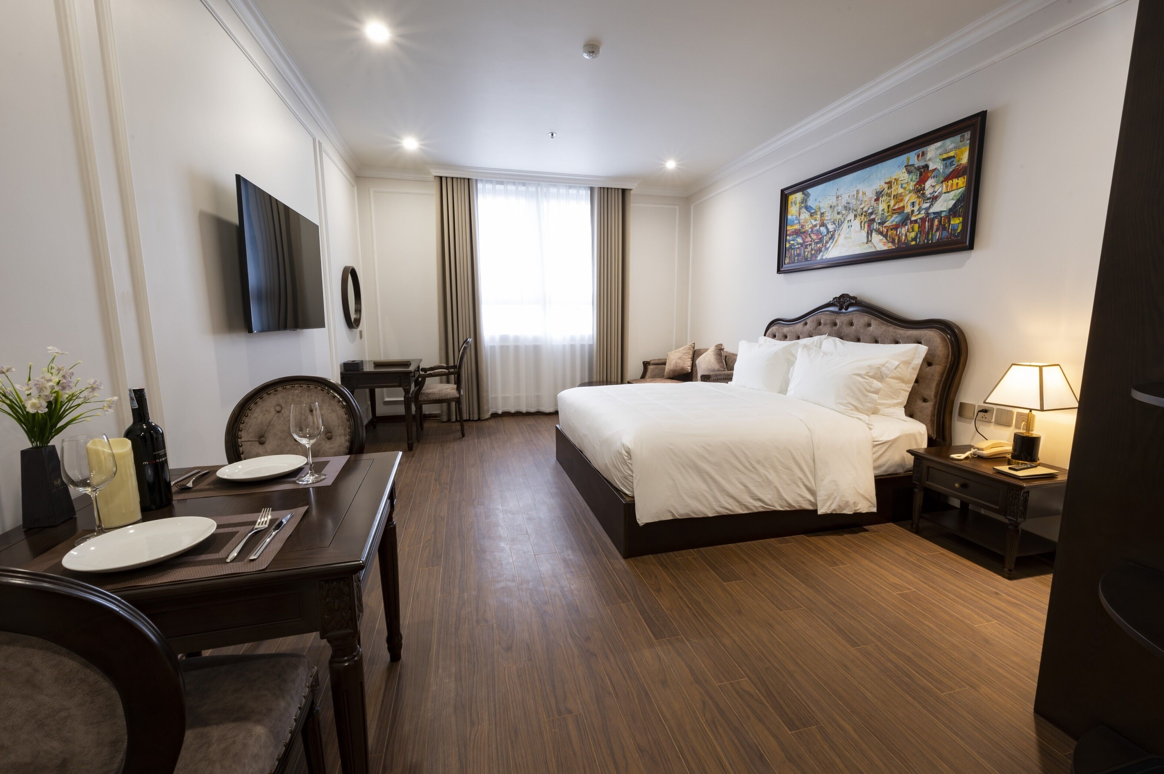 Ein Hotelzimmer mit einem großen Bett, einem Schreibtisch mit Stuhl, einem Esstisch mit Stühlen, einem Fernseher und einem Bild an der Wand.