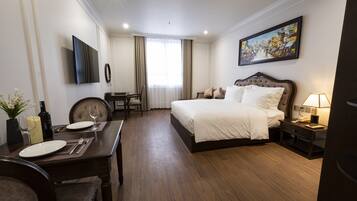 A hotel room with a large bed, a desk with a chair, a dining table with chairs, a TV, and a painting on the wall.