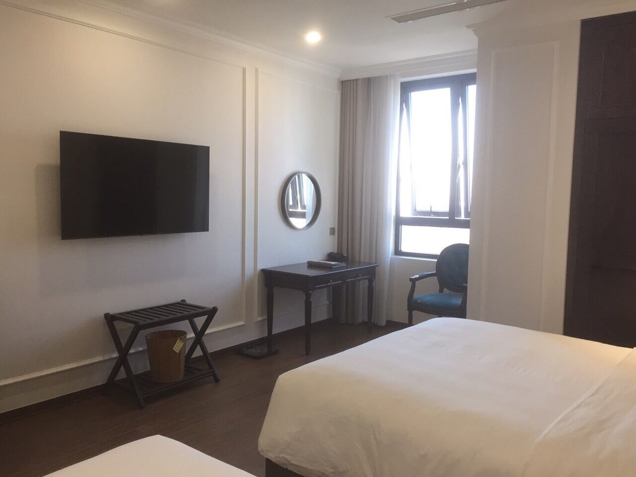 A hotel room with two beds, a flat-screen TV mounted on the wall, a small desk with a chair, a window with curtains, and a waste basket.