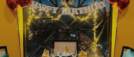 Ett festligt dukat matbord för en födelsedagsfirande, med en banderoll med texten ”Grattis på födelsedagen”, hjärtformade ballonger och ett ljus.