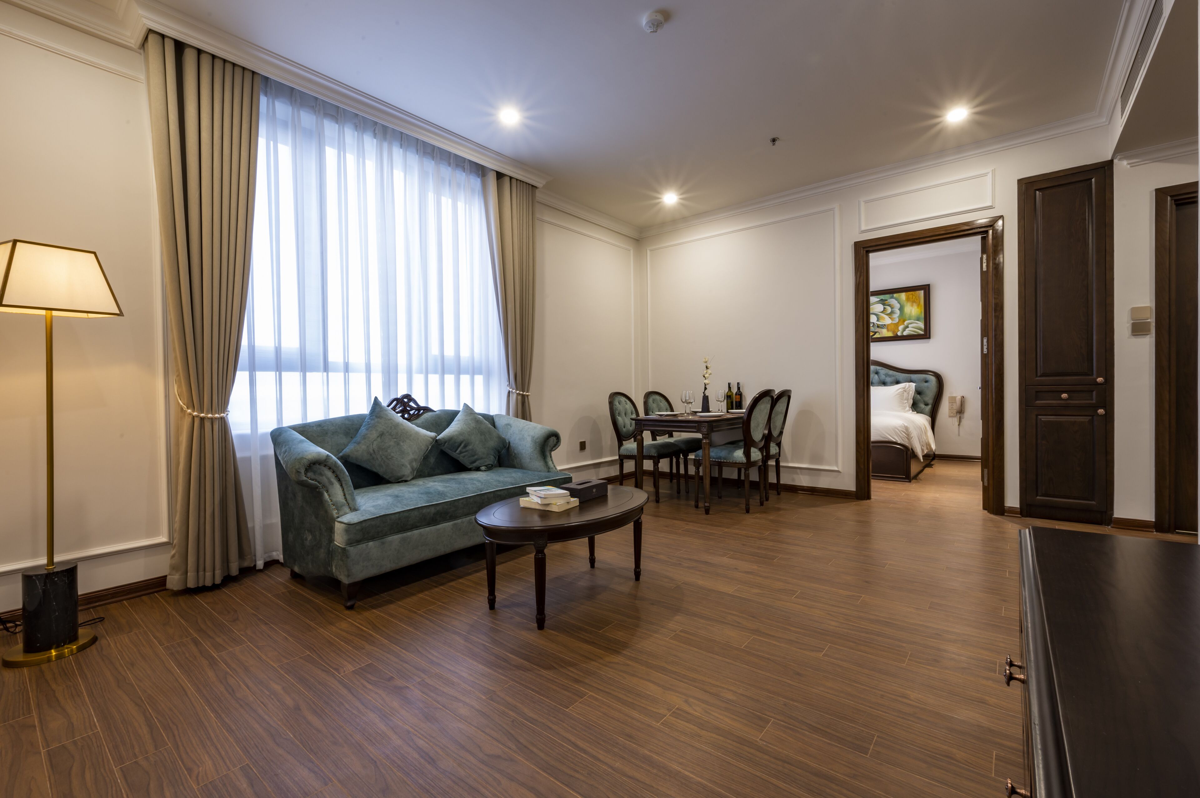 A spacious living room with a sofa, coffee table, and dining area. There is a large window with curtains, a lamp, and a door leading to another room.