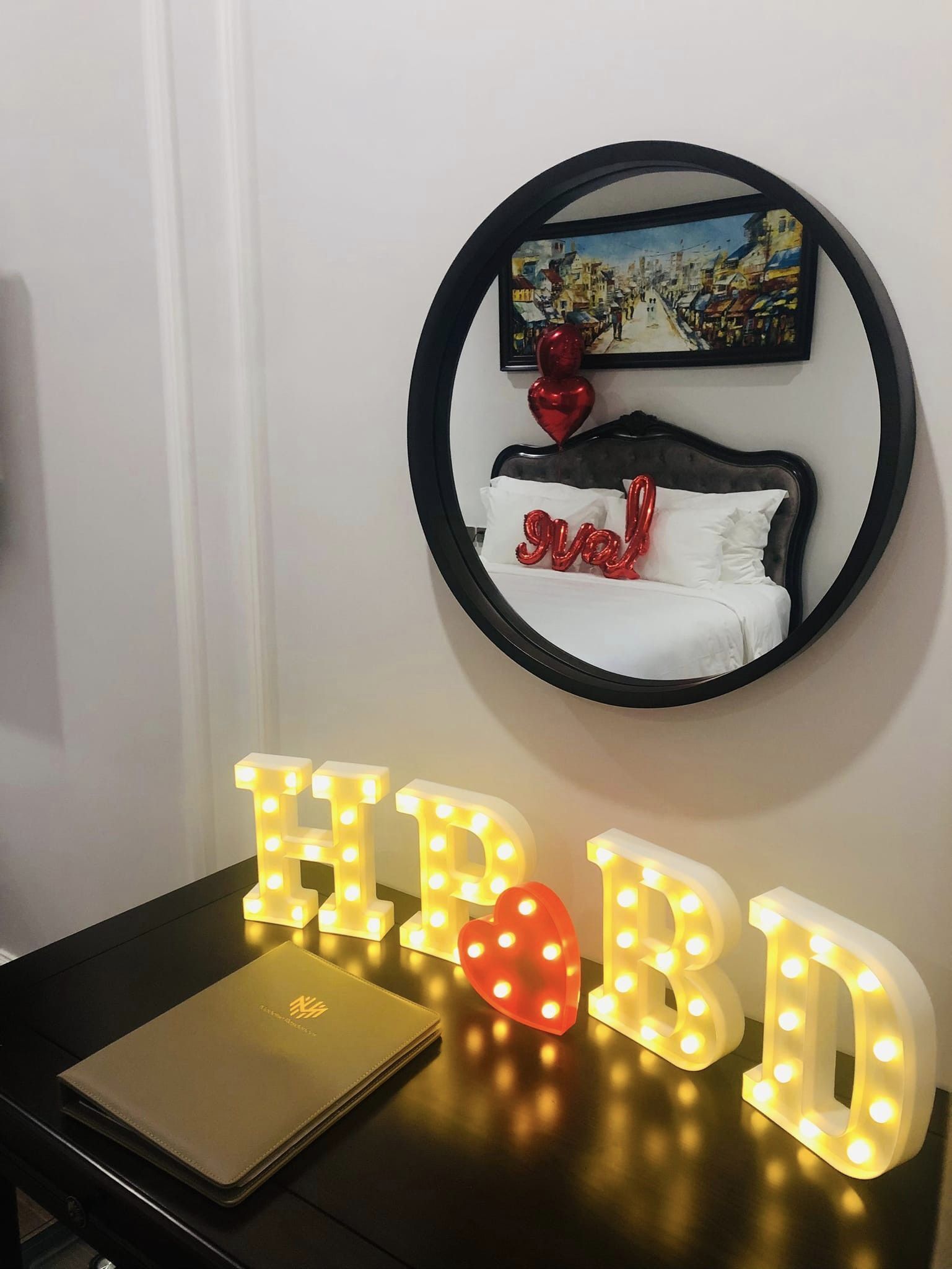 A hotel room with a bed, a round mirror with a painting, and illuminated letters spelling "HBD" on a table.