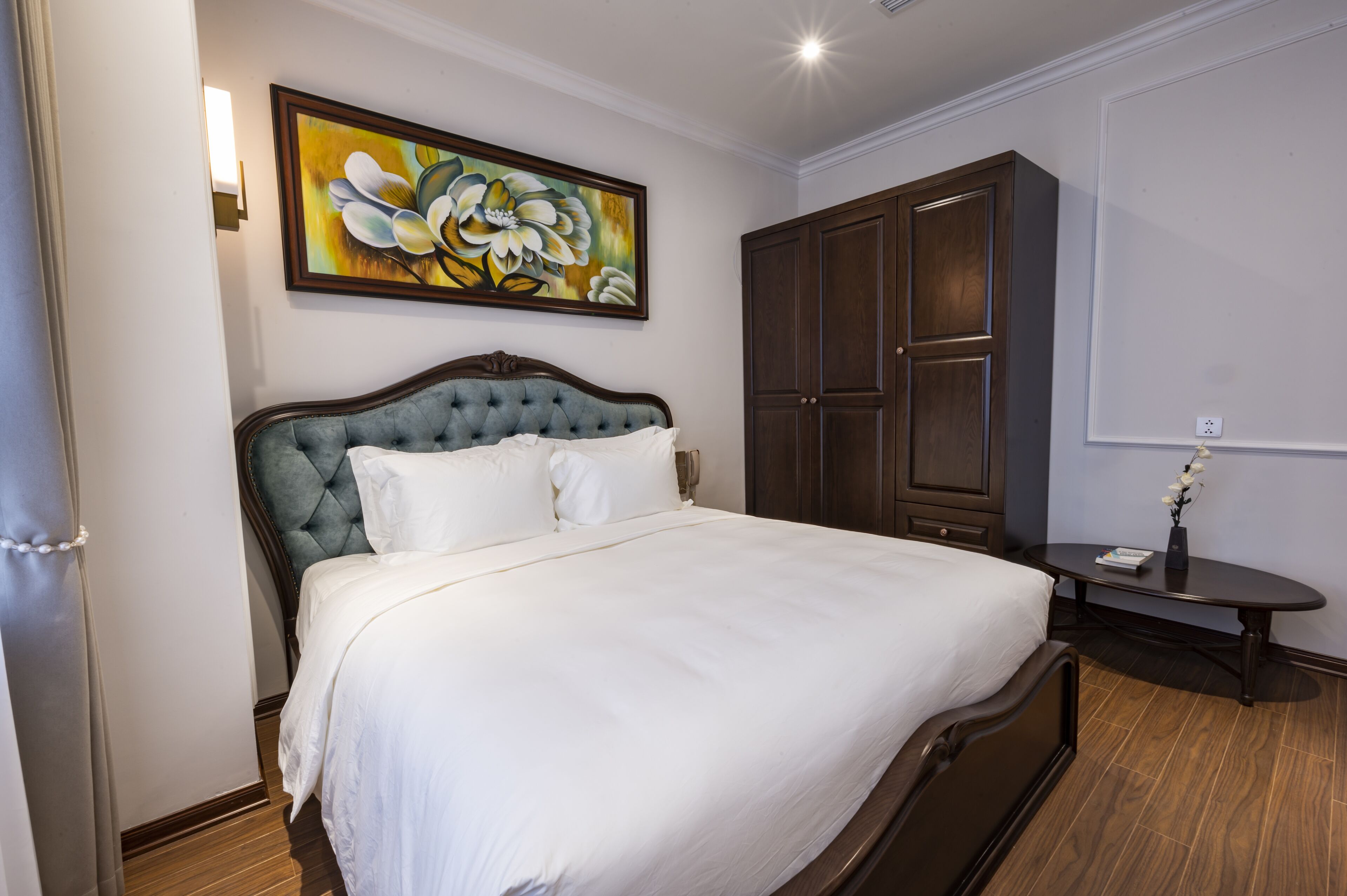 A neatly made bed with a tufted headboard, a large dark wooden wardrobe, and a painting above the bed.