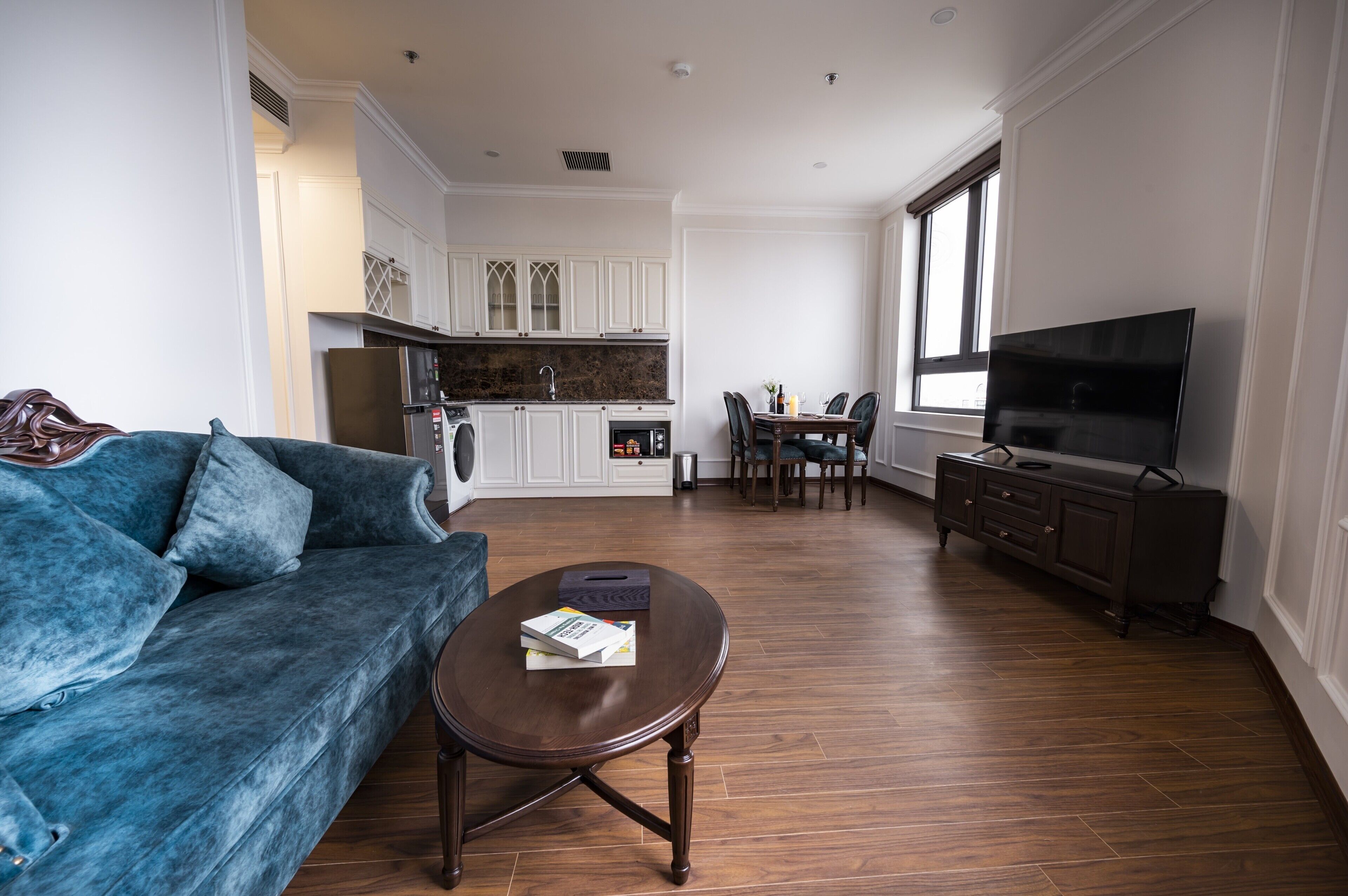 A living room with a blue sofa, a coffee table, a dining area with a table and chairs, a kitchen with white cabinets, and a flat-screen TV on a cabinet.