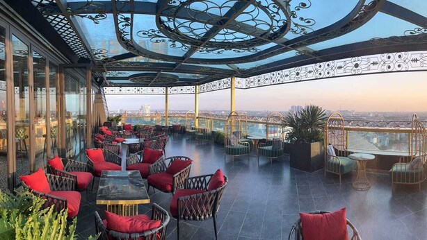 A rooftop terrace with wicker furniture, a glass balustrade, and a city view at sunset.