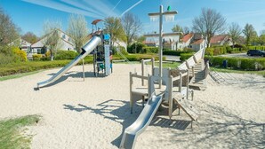 Une aire de jeux avec des toboggans et des structures d’escalade, entourée d’une zone sablonneuse et bordée de maisons d’habitation en arrière-plan.
