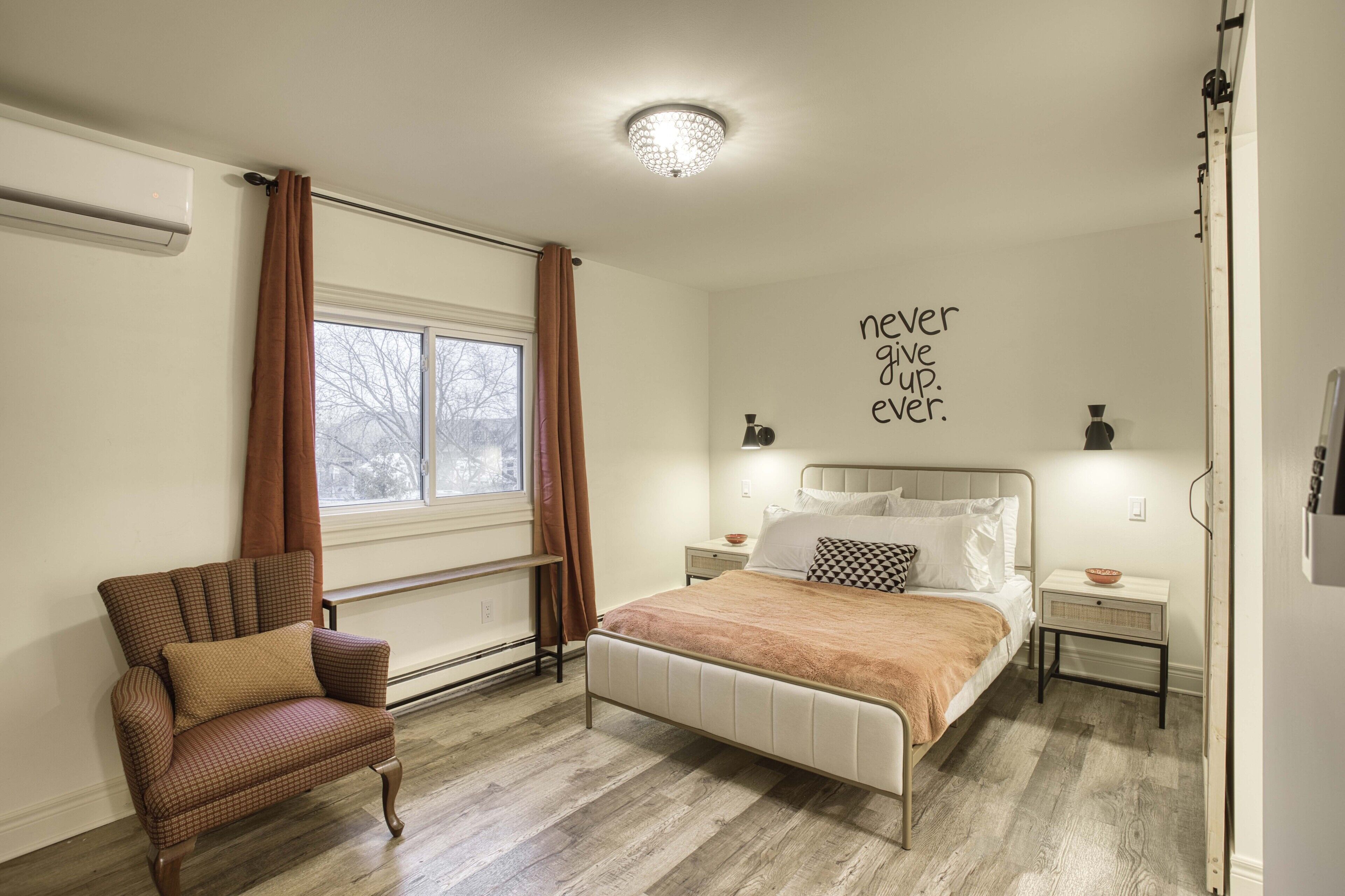Um quarto com cama, uma cadeira, uma janela com cortinas, uma parede com uma frase motivacional e uma luminária no teto.