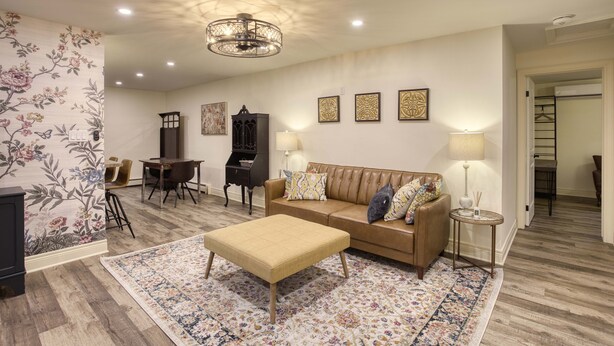 A living room with a sofa, ottoman, and a patterned rug. There is a dining table with chairs, a cabinet, and wall-mounted lamps. - Great for Couples or Family in Niagara Wine Country (Lincoln)