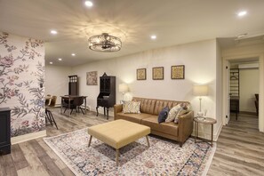 A living room with a sofa, ottoman, and a patterned rug. There is a dining table with chairs, a cabinet, and wall-mounted lamps. - Great for Couples or Family in Niagara Wine Country (Lincoln)