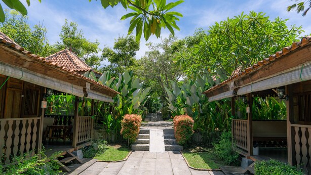 A courtyard with a central pathway leading to a staircase, surrounded by lush greenery and traditional wooden structures with tiled roofs. - Bali Desa Asri (Keroboken Kelod)