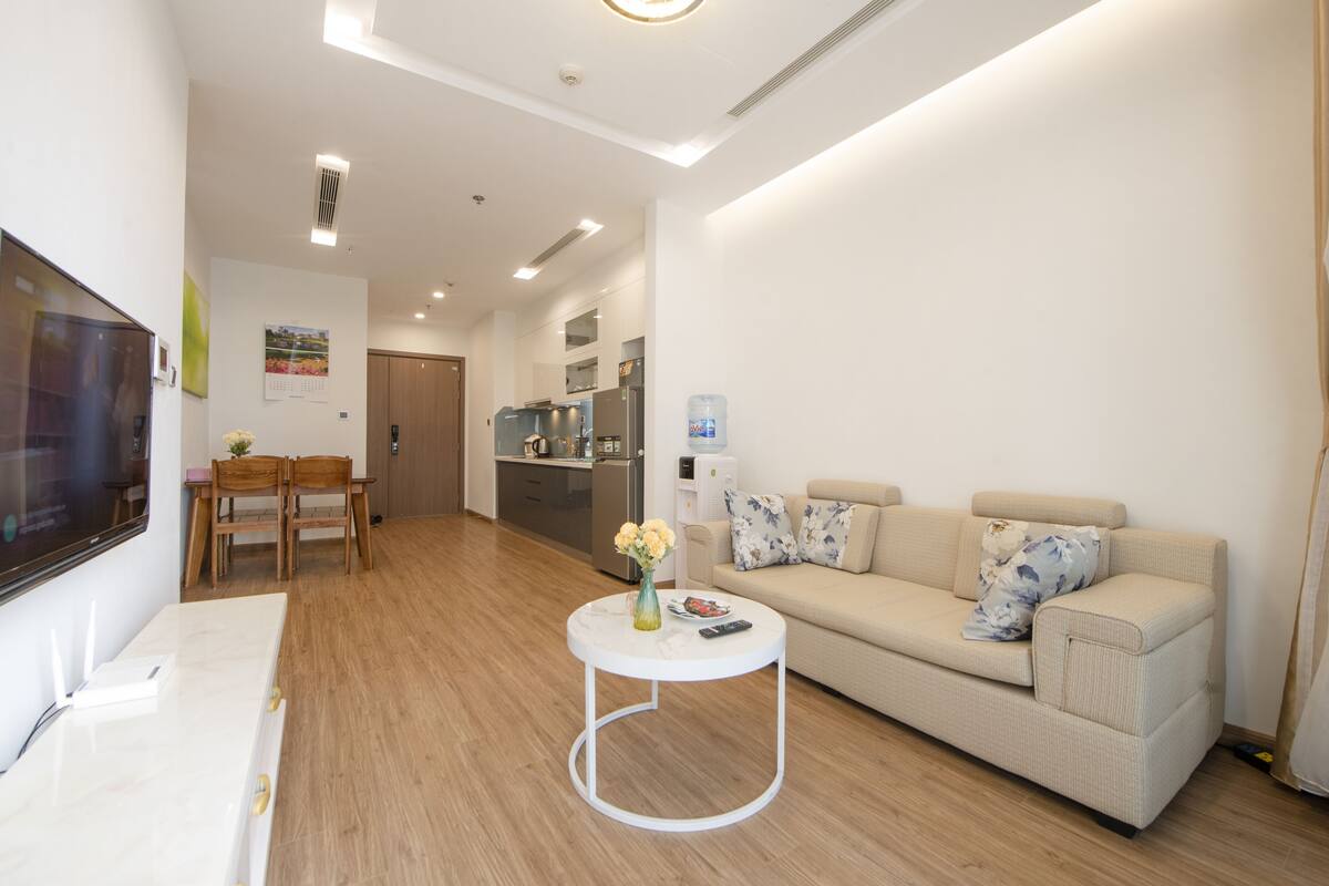Un salon moderne avec un canapé, une table basse, un téléviseur et un coin cuisine à l’arrière-plan.