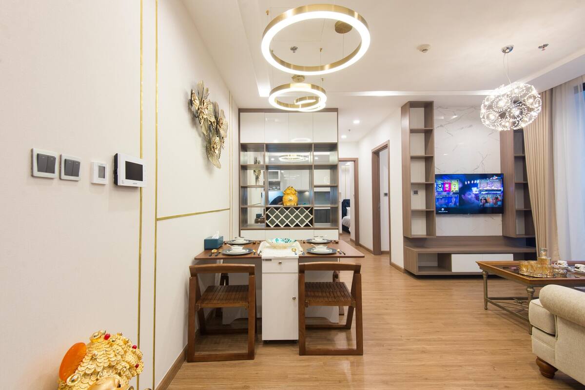 Un salon moderne avec un plancher en bois, un coin repas avec une petite table et des chaises, un coin cuisine avec un réfrigérateur et un évier, et un téléviseur fixé au mur.
