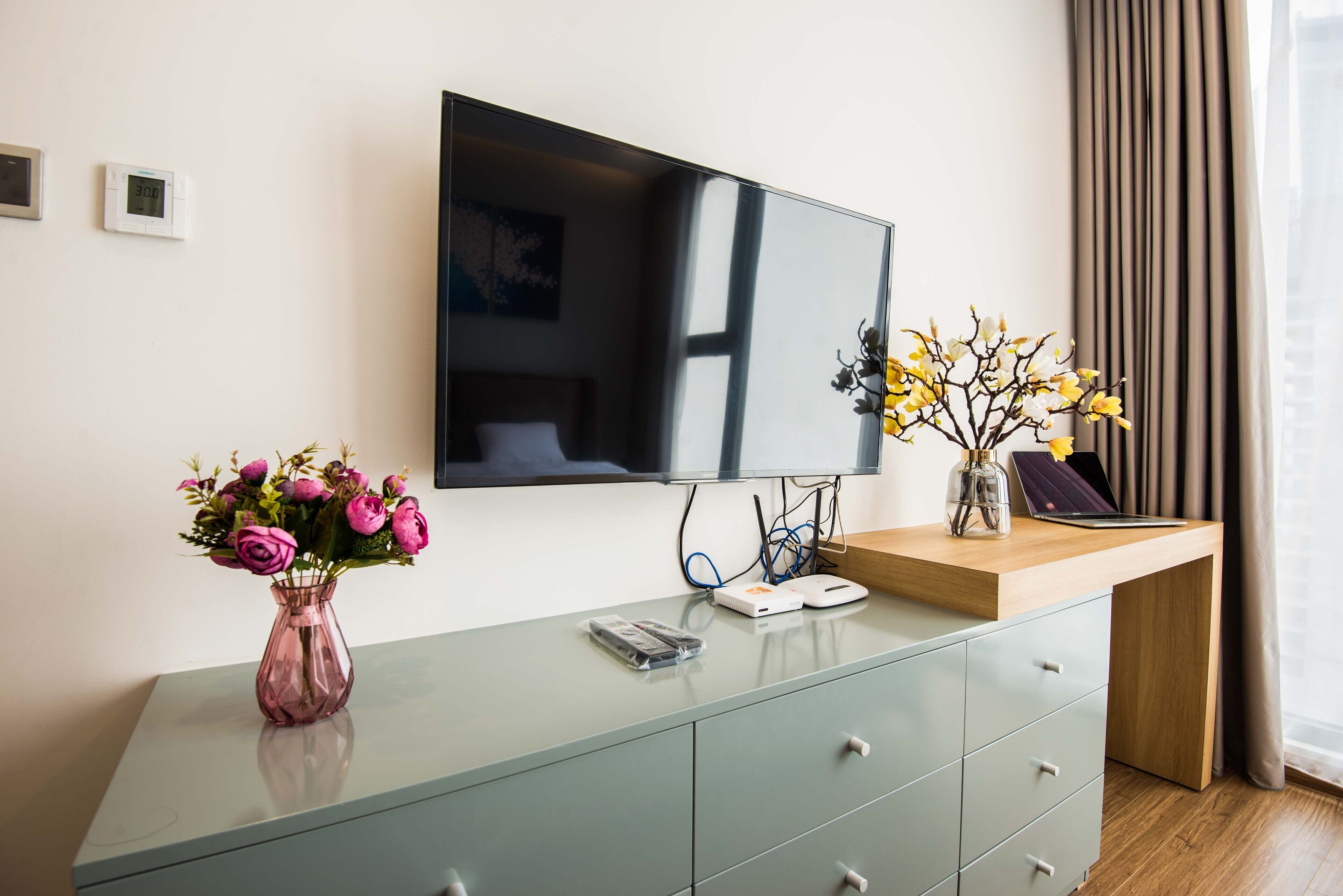 A modern hotel room with a flat-screen TV mounted on the wall, a glass desk with a vase of flowers, and a wooden side table with a laptop and a decorative vase.