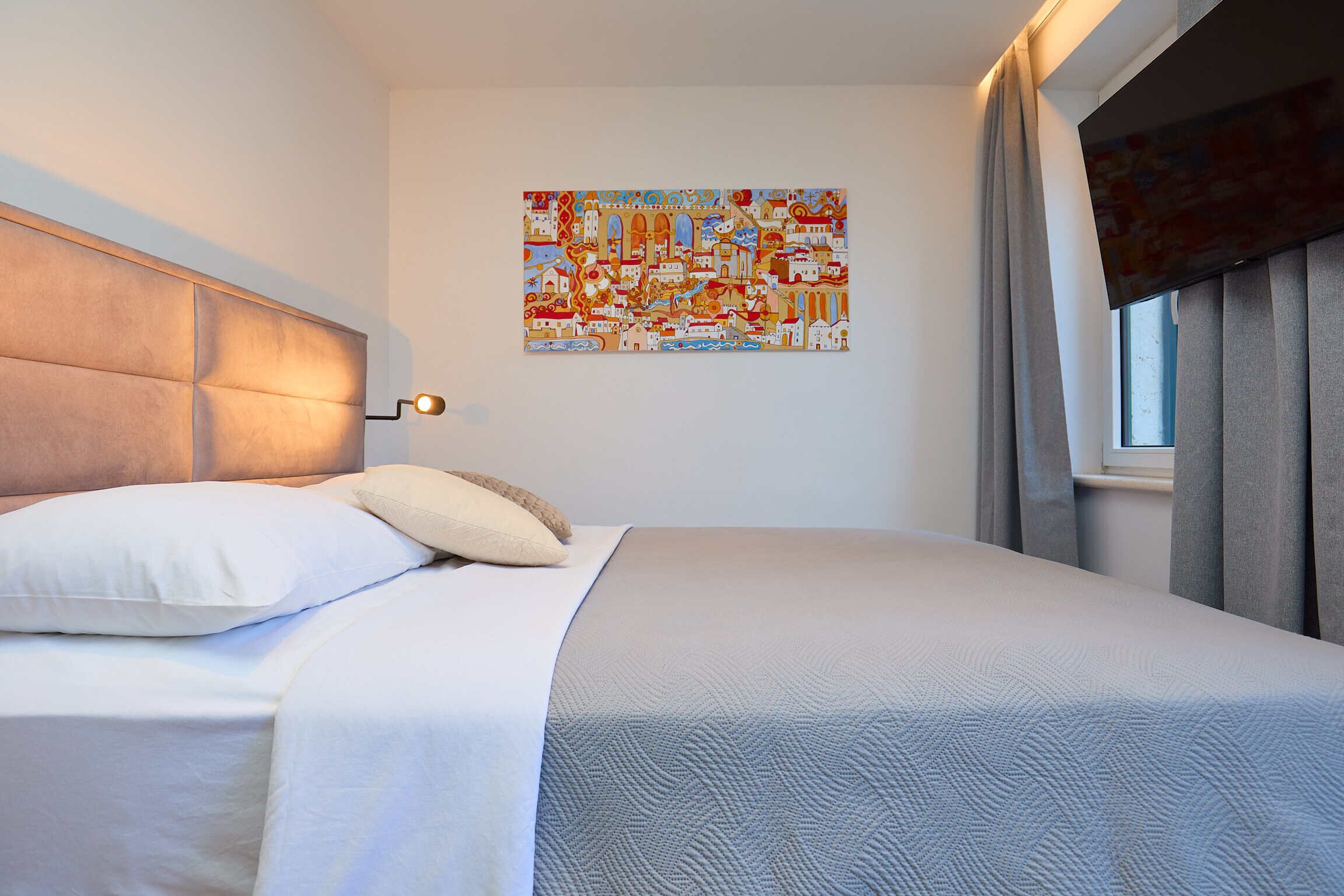 A neatly made bed with white linens and a headboard, a wall-mounted artwork depicting a colorful scene, and a window with curtains.