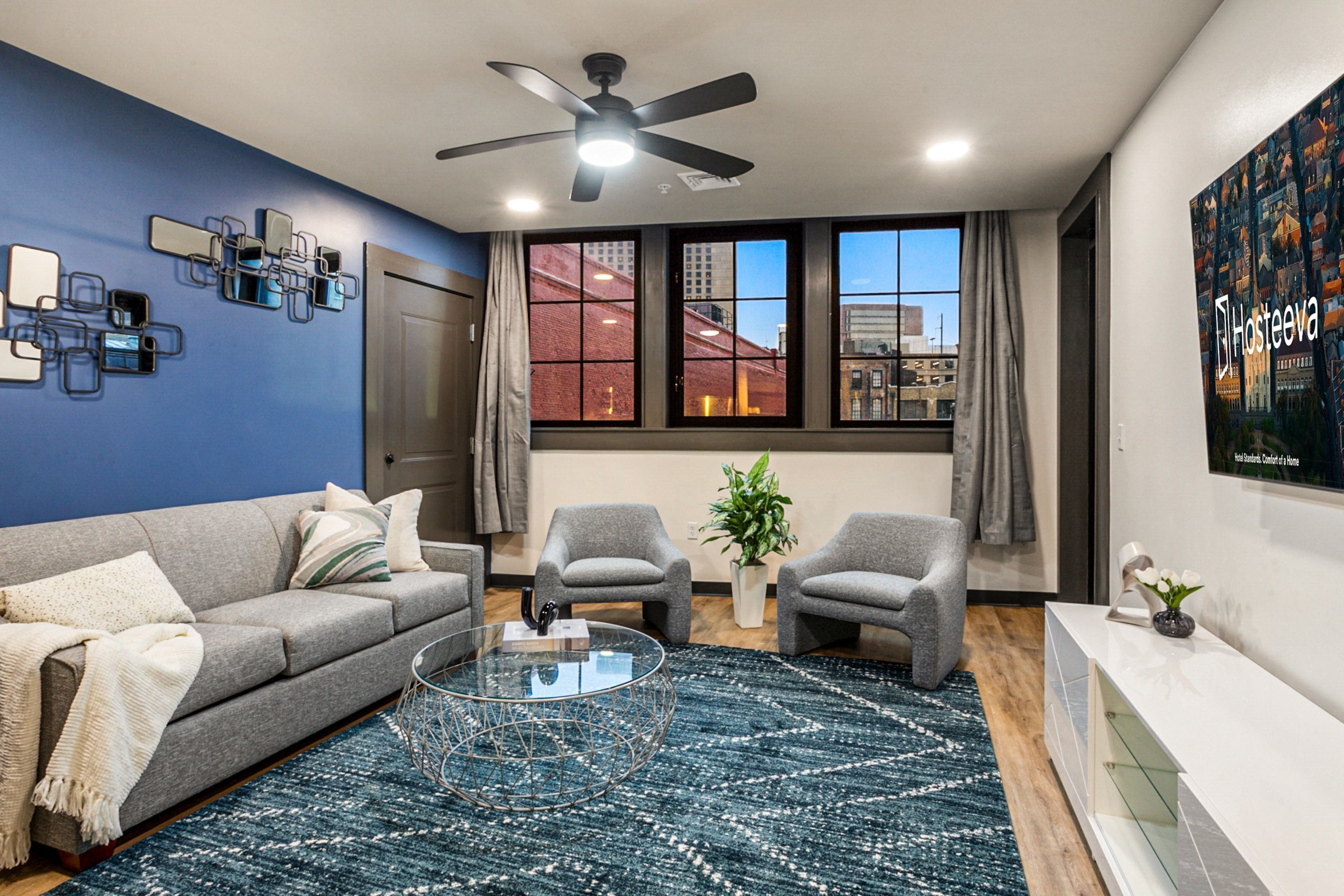A modern living room with a grey sofa, two armchairs, a glass coffee table, and a blue patterned rug. There is a ceiling fan, a large window with curtains, and a poster on the wall.