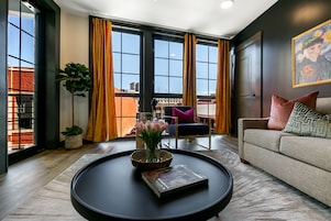 A modern living room with a large window, a black coffee table with a book and flowers, a sofa with cushions, and a painting on the wall.