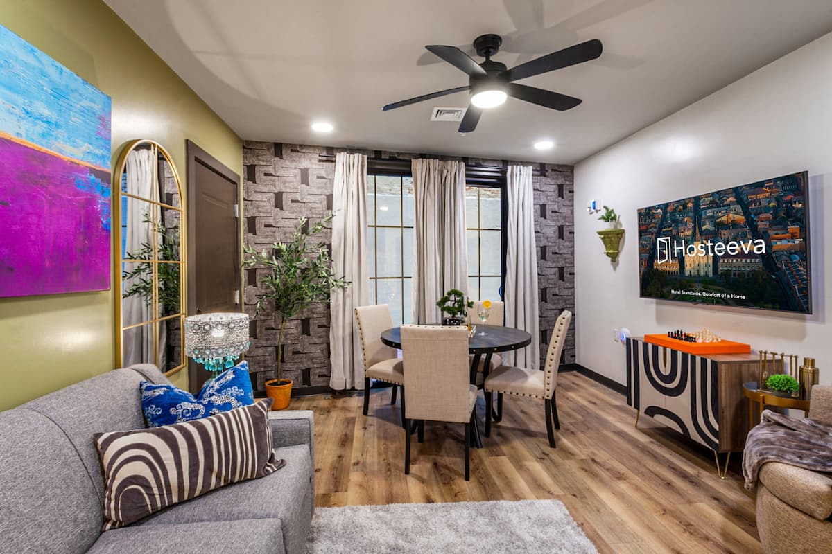 Un salon moderne comprenant un coin repas, un ventilateur de plafond, un téléviseur fixé au mur et une grande fenêtre avec des rideaux.