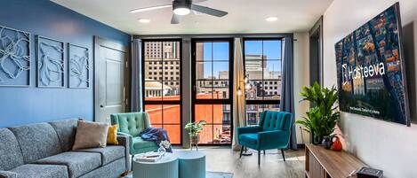A modern hotel room with a grey sofa, teal armchairs, a wooden console table, and a large window offering city views.