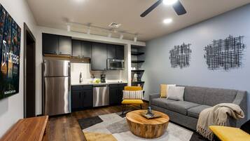 A modern kitchen with stainless steel appliances, a dining area with a round wooden table, and a grey sofa.