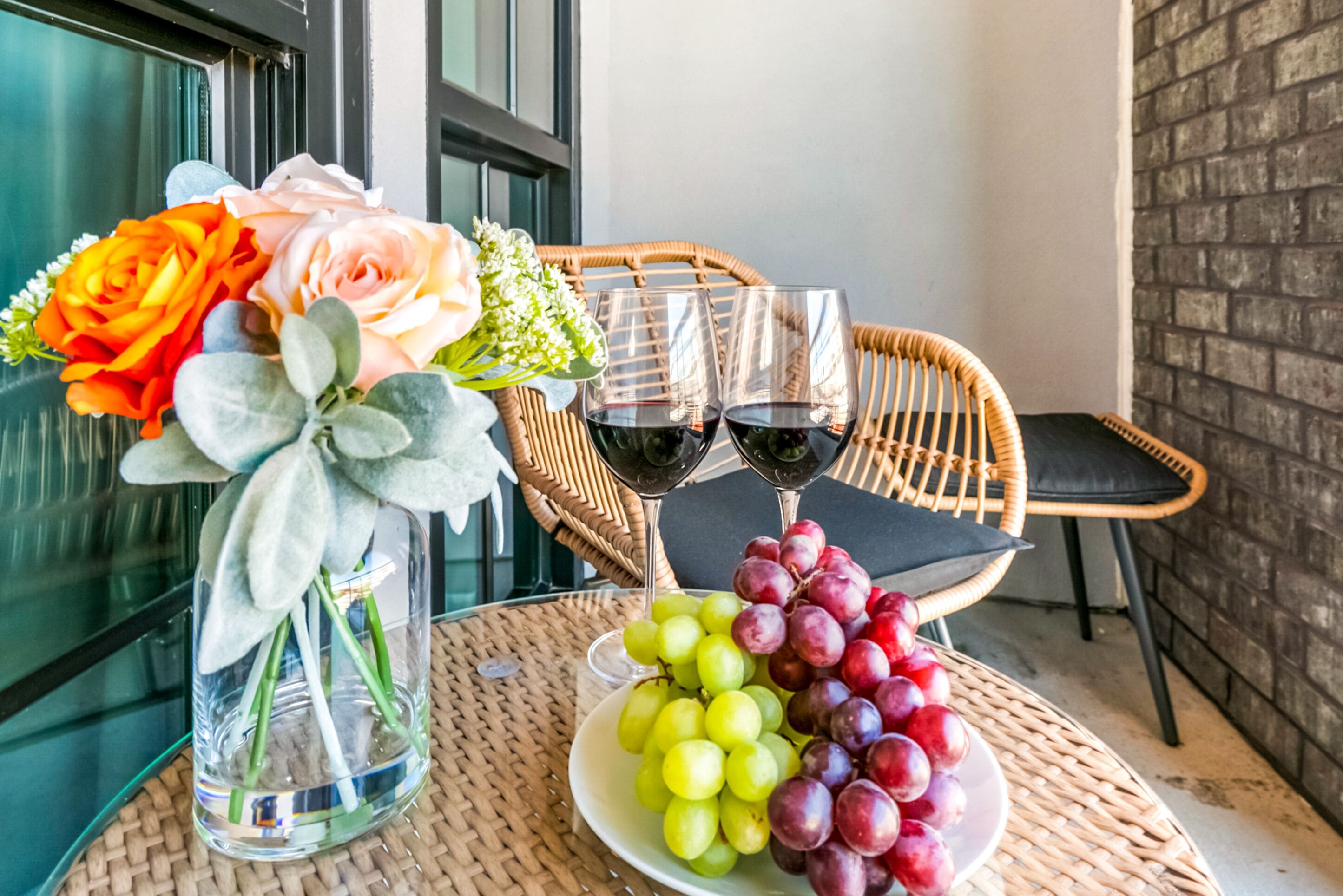 Une table en osier sur laquelle se trouve un vase en verre rempli de fleurs, deux verres à vin et une assiette de raisins.
