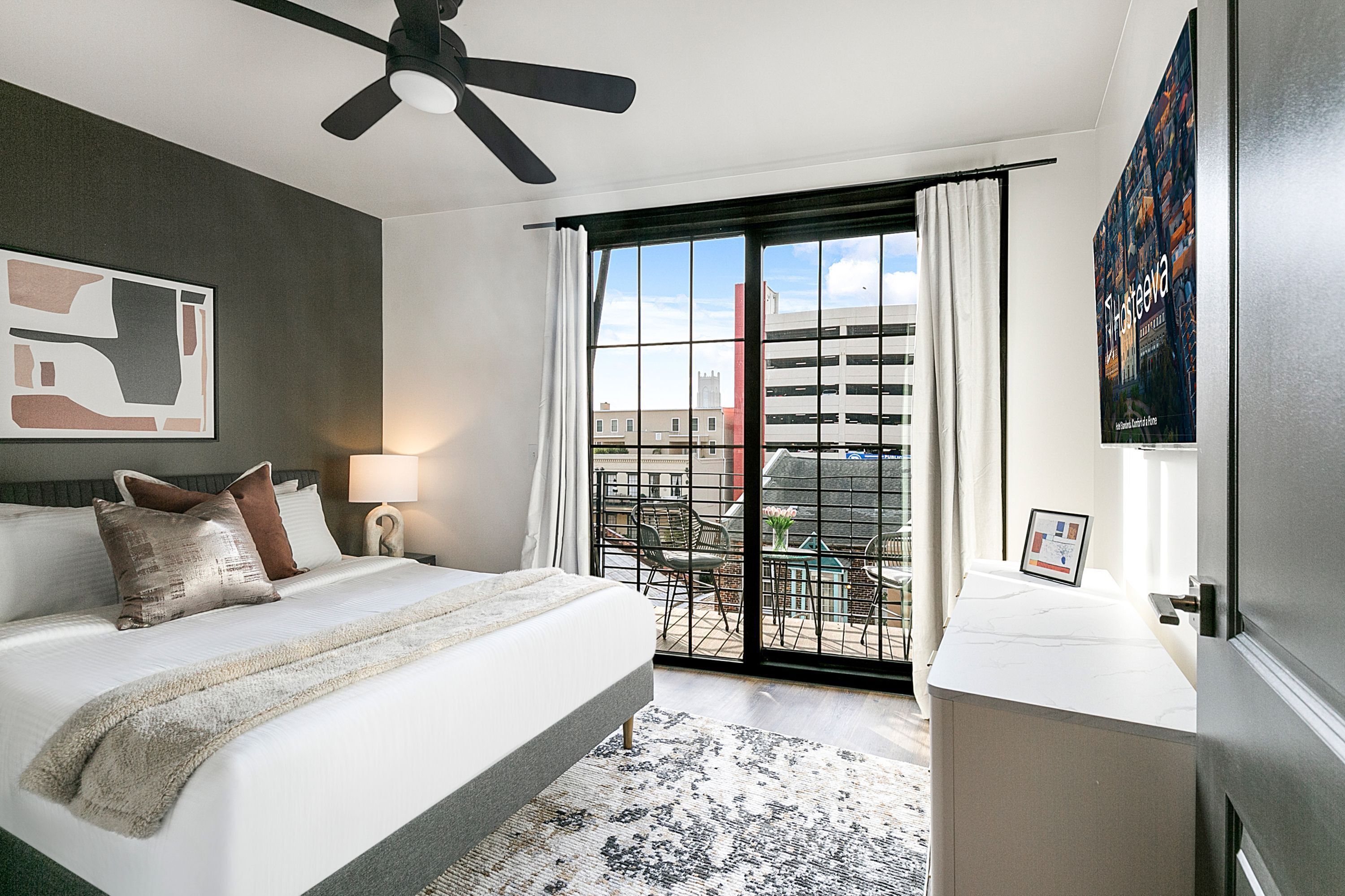 Habitación de hotel con cama, ventilador de techo, ventana grande con cortinas y vista a edificios, además de una obra de arte colgada en la pared.