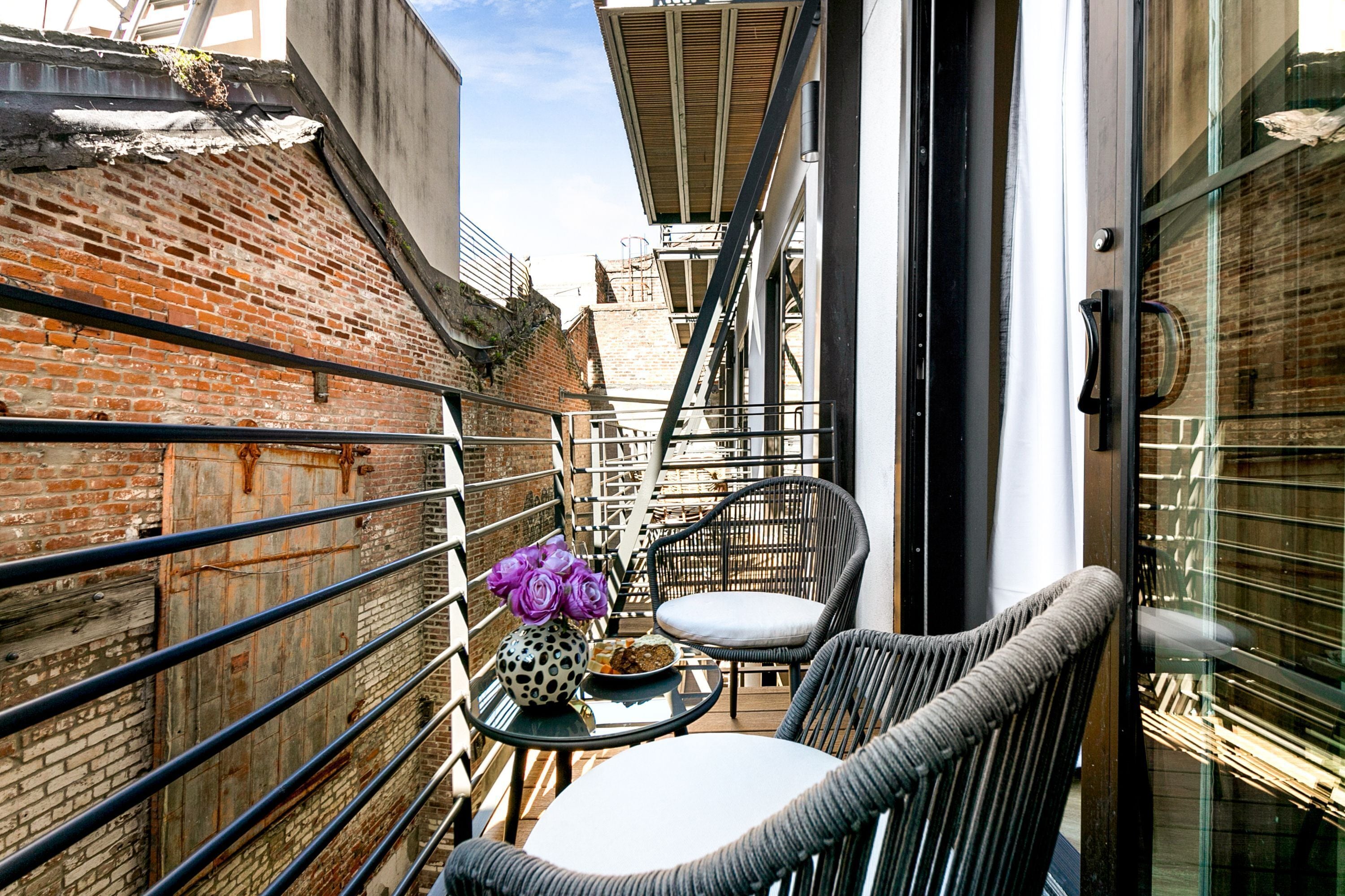 A narrow outdoor balcony with wicker chairs and a small table, a vase of purple flowers, and a brick wall.