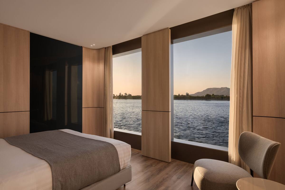 A modern hotel room with a large bed, a chair, and a view of a body of water through large windows.