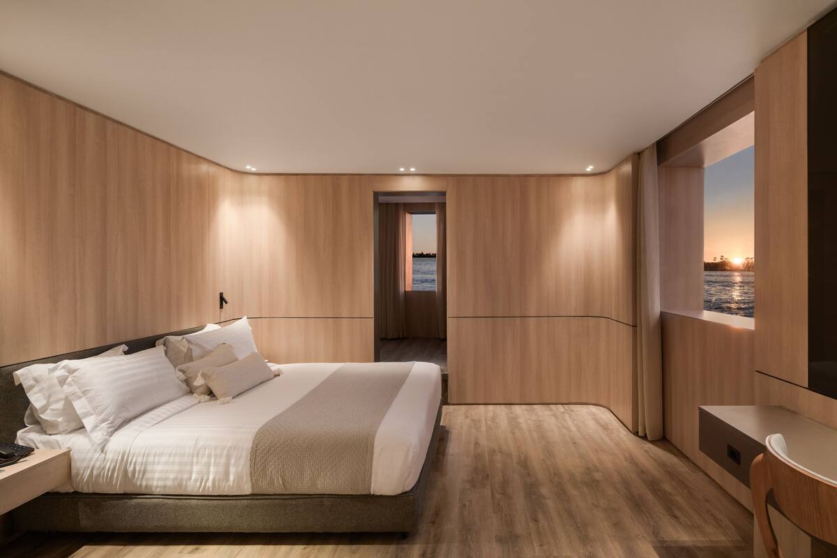 A modern bedroom with a large bed, wooden paneling, a desk, and a view of the sea.