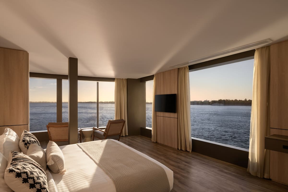 A modern hotel room with a large bed, a TV, a chair, and a view of the water.