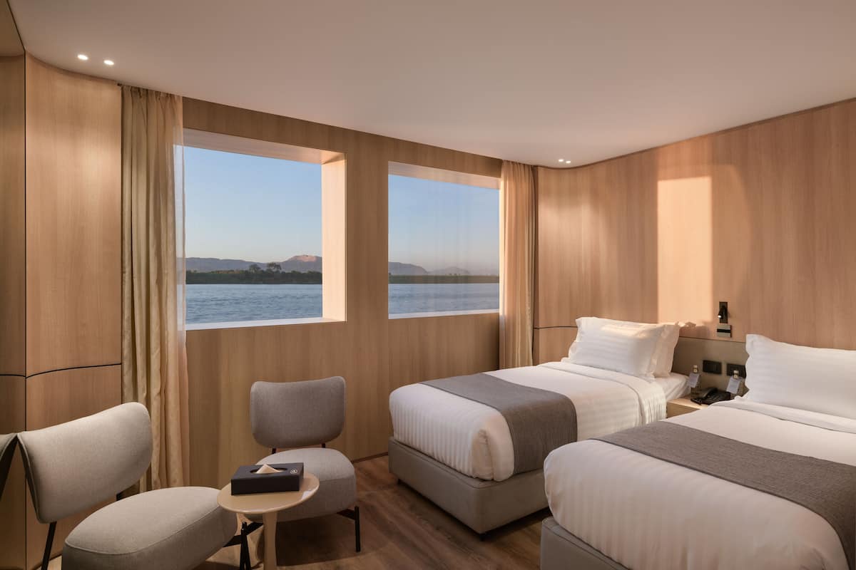 A hotel room with two beds, a small table, a chair, and large windows offering a view of the sea.