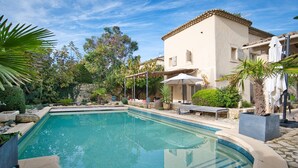 Une piscine bordée de pierres, entourée d’un jardin où poussent différentes espèces de plantes, et un bâtiment surmonté d’un toit en tuiles.