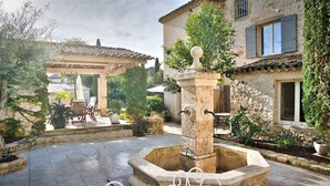 Une cour avec une fontaine en pierre, des sièges en plein air et un bâtiment aux murs et aux volets en pierre.