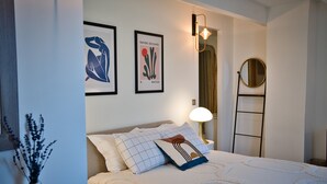 Une chambre à coucher bien rangée, avec un lit, des oreillers, une lampe de chevet, un miroir fixé au mur et deux œuvres d’art encadrées.