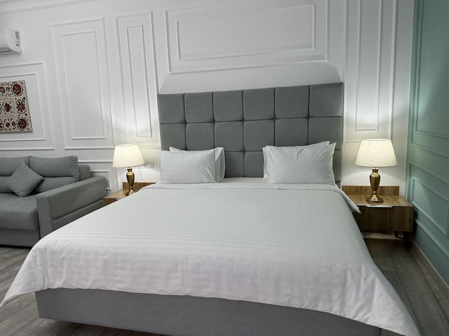 A neatly arranged bedroom with a large bed, two bedside tables with lamps, a grey sofa, and a decorative wall piece.