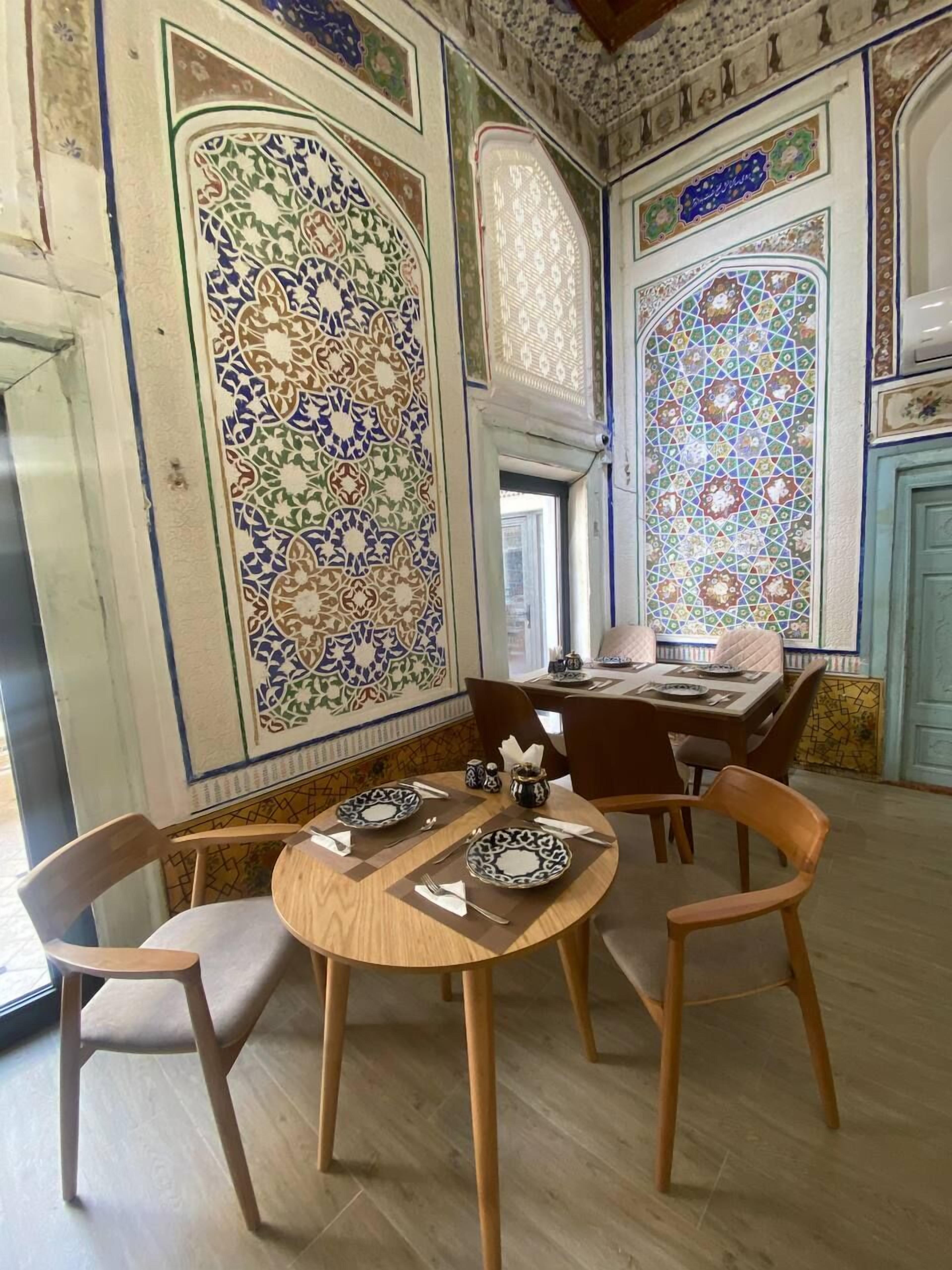 Un comedor con una mesa de madera redonda dispuesta para dos personas, rodeada de sillas de madera. Las paredes están decoradas con intrincados diseños de azulejos.