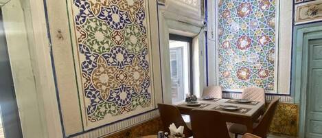 A dining area with a round wooden table set for two, surrounded by wooden chairs. The walls are adorned with intricate tile patterns.