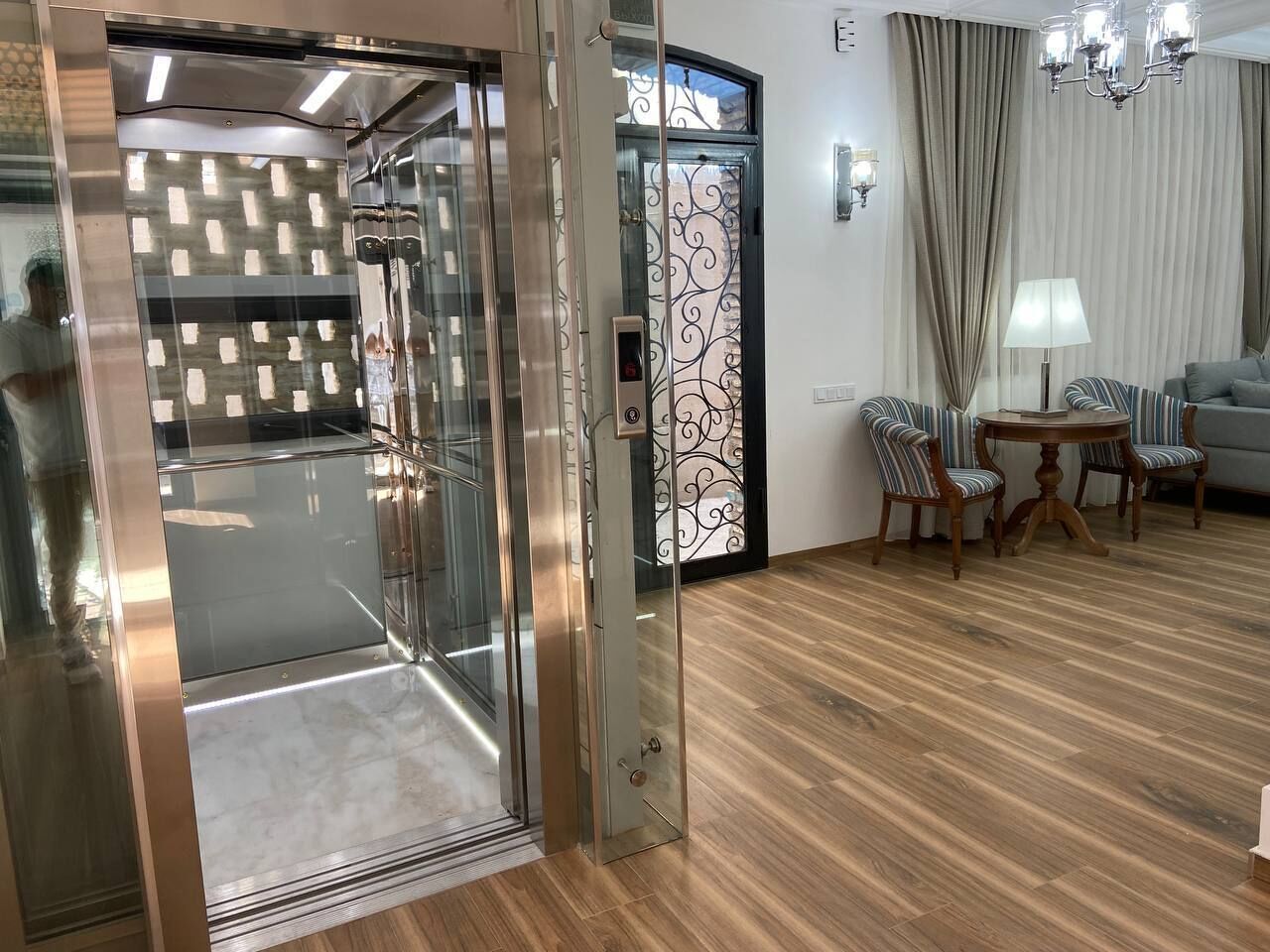 A modern room with a glass elevator, wooden flooring, a sofa set, and a table with chairs.