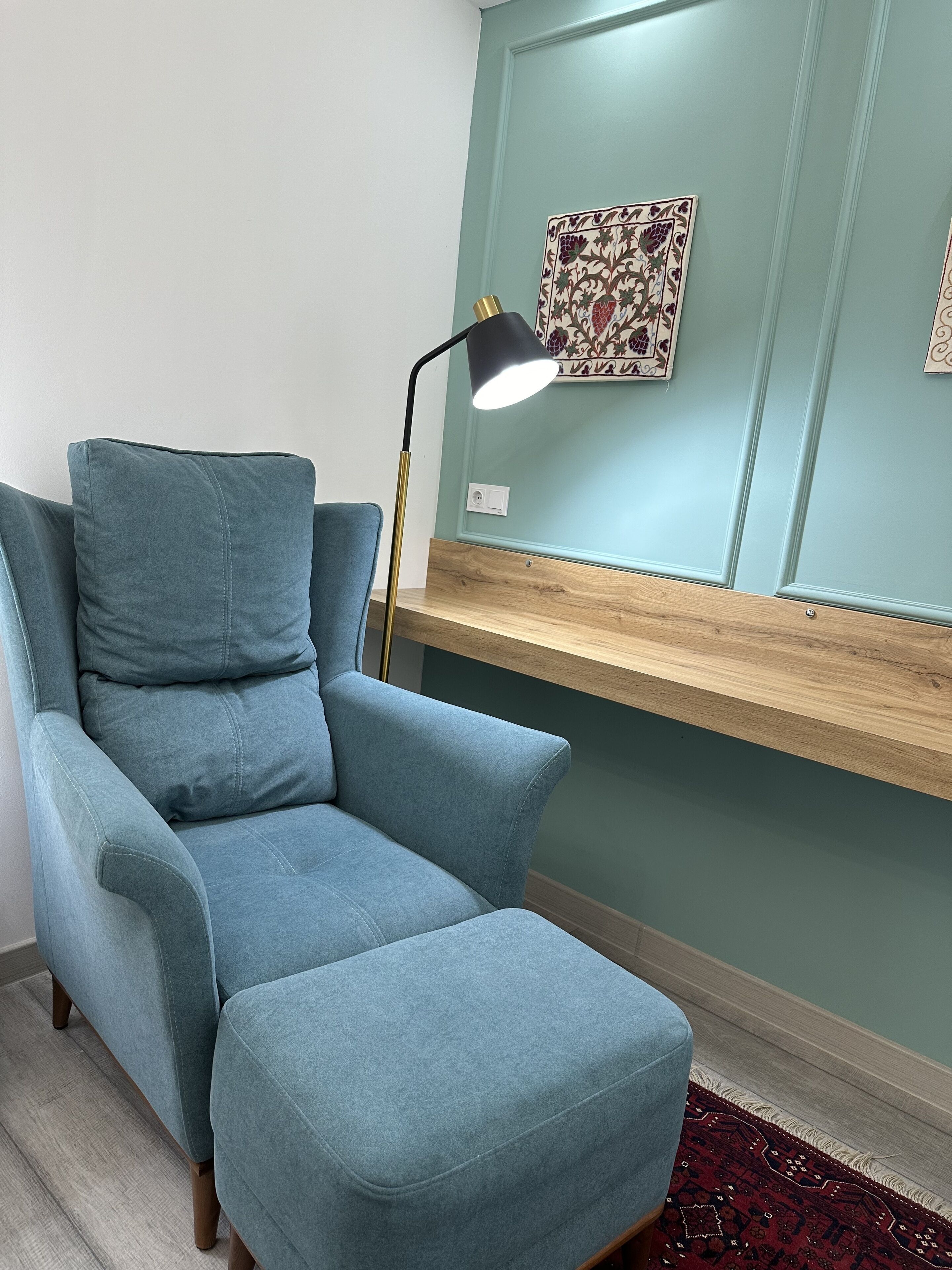 A room with a blue armchair, a desk with a light fixture, and a patterned wall hanging.
