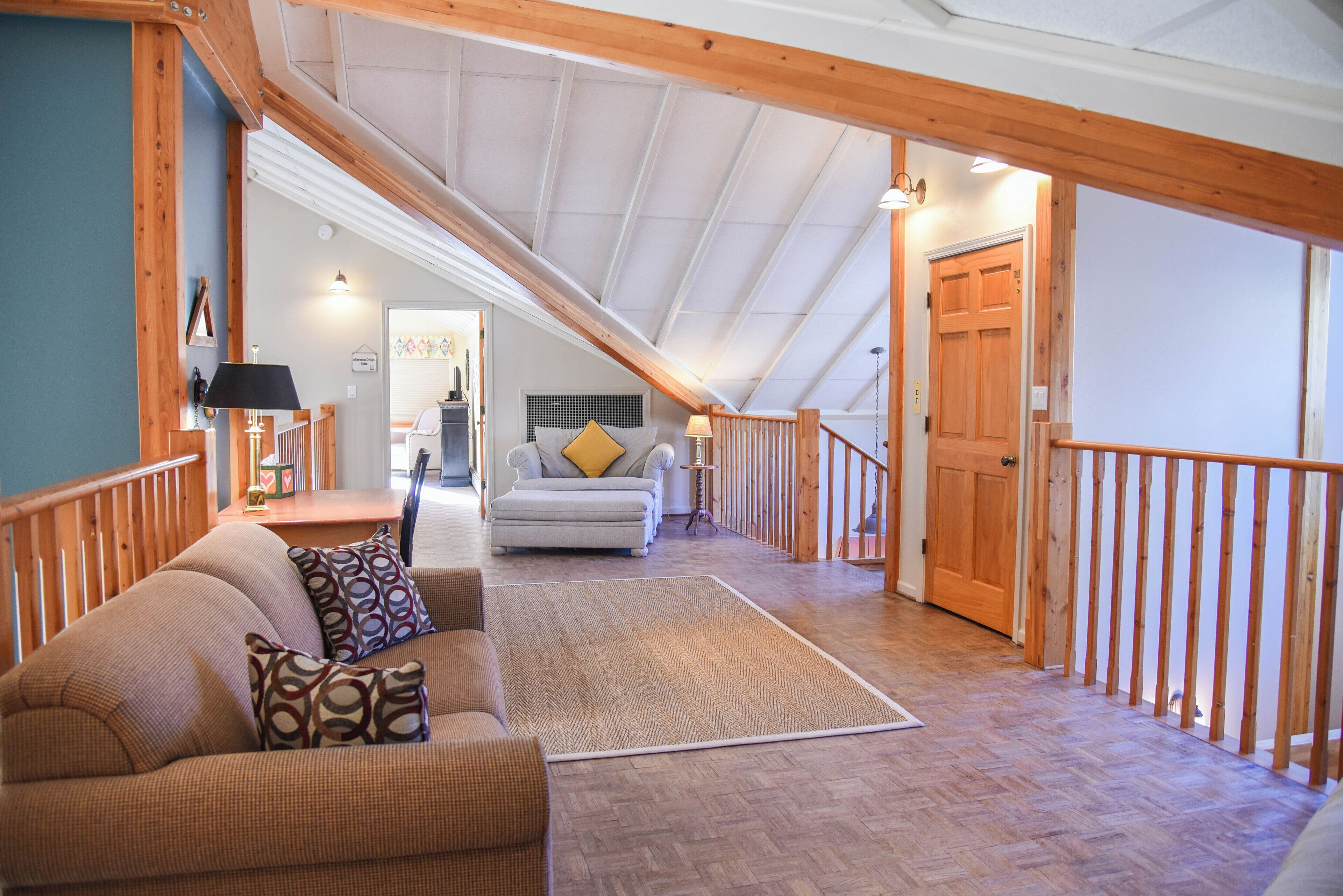 Une chambre spacieuse aménagée en mezzanine, avec un plafond haut, des poutres en bois, une grande fenêtre et un coin salon confortable avec un canapé et des coussins à motifs.